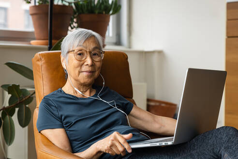Porträt einer älteren Frau, die Kopfhörer trägt, während sie zu Hause einen Laptop benutzt - CAVF94979