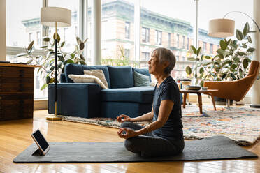 Senior woman meditating while learning through digital tablet in living room at home - CAVF94983