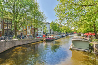Gekräuselter Fluss mit vertäuten Motorschiffen und Hausbooten zwischen Bäumen und Gebäudefassaden im Sonnenlicht im Hafen von Amsterdam - ADSF31360