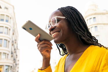 Positive African American female with black hair in sunglasses recording voice message on modern smartphone while strolling through the city streets - ADSF31368