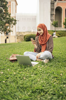 Muslimische Frau, die im Gras sitzt und einen Computer und ein Telefon benutzt - CAVF95012