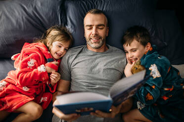 Father reading a book to his children in the master bedroom at night - CAVF95044