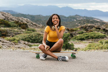 Ganzkörperaufnahme einer fröhlichen ethnischen Frau mit drahtlosen Kopfhörern im Nacken, die auf einem Longboard sitzt und in die Kamera schaut - ADSF31394