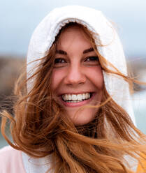 Porträt einer charmanten, fröhlichen, rätselhaften Frau mit fliegendem rotem Haar und Kapuze, die an einem windigen Tag in Galicien in die Kamera schaut - ADSF31395