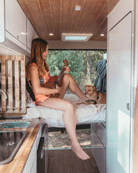 Seitenansicht einer fröhlichen Reisenden, die mit ihrem Hund in einem Van auf dem Bett sitzt und Ukulele spielt, während einer Reise in Spanien - ADSF31396
