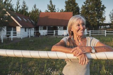 Lächelnde ältere Landarbeiterin lehnt am Zaun eines Bauernhofs - MASF26372