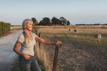 Lächelnde ältere Bäuerin, die am Zaun steht und auf ein Feld blickt - MASF26374