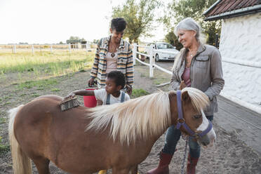 Boy brushing horse while standing with grandmother and mother at farm - MASF26410