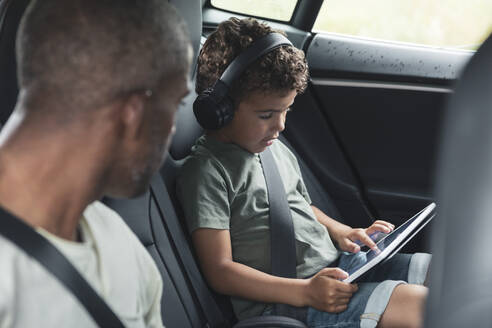 Mann schaut seinem Sohn zu, der im Elektroauto ein Video auf einem digitalen Tablet anschaut - MASF26530