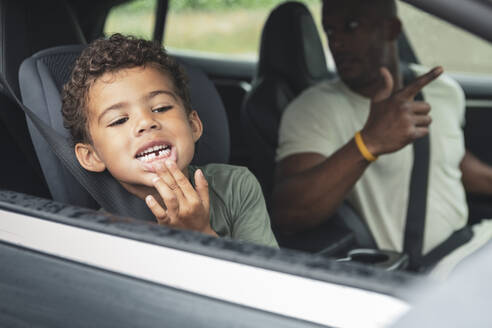 Playful boy showing broken teeth while sitting by father in electric car - MASF26535