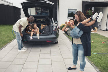 Smiling woman carrying son while man looking at daughter sitting in car trunk - MASF26544