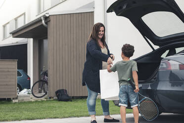 Lächelnde Mutter gibt ihrem Sohn, der neben einem Elektroauto im Vorgarten steht, eine Tasche - MASF26548