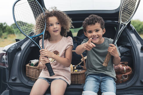 Boy holding tennis racket while pointing by sister sitting in car trunk - MASF26554