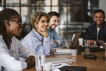 Smiling businesswoman looking away while sitting amidst colleagues at conference table - MASF26610