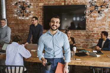 Porträt eines lächelnden Geschäftsmannes mit Kaffeetasse und Akte im Stehen, während Kollegen im Hintergrund diskutieren - MASF26612