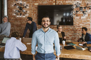 Portrait of happy male professional standing while colleagues discussing in background - MASF26614