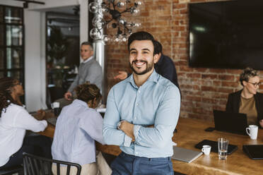 Portrait of smiling male entrepreneur standing with arms crossed while colleagues discussing in background - MASF26615