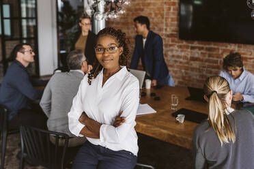 Portrait of businesswoman standing with arms crossed while colleagues discussing in background - MASF26621