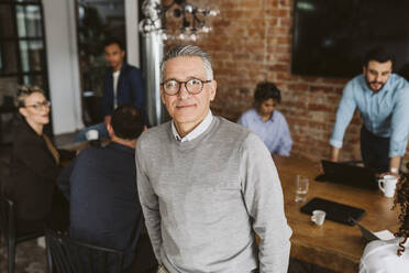 Portrait of mature male entrepreneur standing while colleagues discussing in background - MASF26623