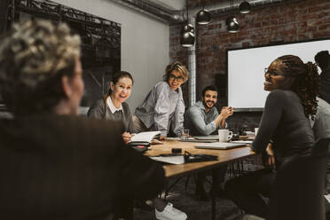 Smiling male and female professionals looking at mature entrepreneur while sitting at conference table - MASF26634