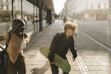 Jugendlicher mit Skateboard, der lachend neben einer Freundin auf einem Fußweg in der Stadt steht - MASF26711