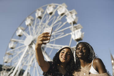 Multirassische Freundinnen machen ein Selfie mit ihrem Smartphone vor einem Riesenrad an einem sonnigen Tag - MASF26713