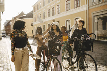 Friends with bicycles looking at female friend holding skateboard in city - MASF26733