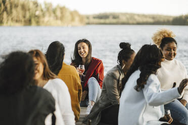 Lächelnde multirassische Freundinnen sitzen auf einem Steg am See - MASF26784