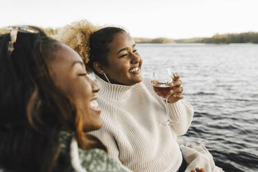 Smiling woman enjoying wine with female friend at lakeshore - MASF26785