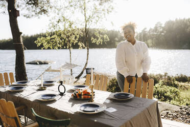 Happy woman standing at dining table against lake - MASF26828