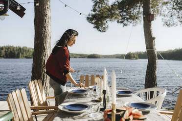 Young woman setting table by lake on sunny day - MASF26831