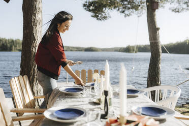 Smiling young woman setting table by lake on sunny day - MASF26832