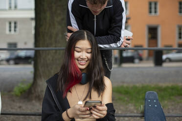 Teenager, der eine lächelnde Freundin betrachtet, die ein Smartphone im Park benutzt - MASF26866