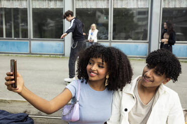 Teenager-Mädchen nimmt Selfie mit männlichen Freund durch Smartphone beim Sitzen auf Bank - MASF26928