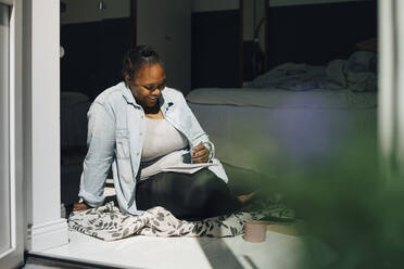 Smiling woman writing on book in bedroom doorway at home - MASF26969