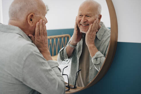 Lächelnder älterer Mann, der seine Haut berührt, während er in den Spiegel schaut - MASF26989
