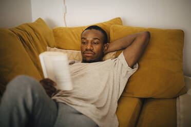 Mid adult man reading book while lying on sofa at home - MASF26996