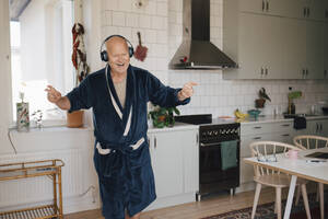 Cheerful senior man wearing headphones dancing in kitchen at home - MASF27017