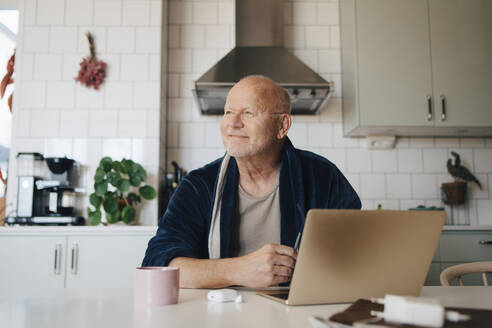 Lächelnder älterer Mann mit Laptop, der wegschaut, während er in der Küche zu Hause sitzt - MASF27050
