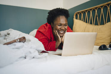 Smiling woman watching on laptop while lying on bed at home - MASF27059