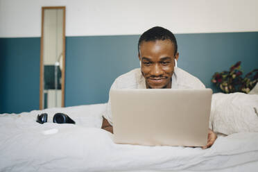 Lächelnder mittelgroßer erwachsener Mann, der einen Laptop benutzt, während er zu Hause auf dem Bett liegt - MASF27073