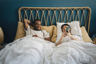 Lächelnde Freundin teilt ihr Smartphone mit ihrem Freund, während sie auf dem Bett liegt - MASF27076