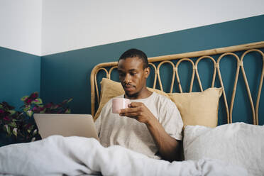 Mittlerer erwachsener Mann arbeitet am Laptop und trinkt Kaffee im Bett - MASF27077