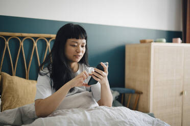 Junge Frau mit Smartphone auf dem Bett im Schlafzimmer - MASF27079