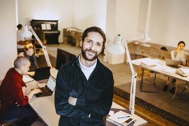 Portrait of smiling male programmer standing with arms crossed while colleagues working at desk in office - MASF27106