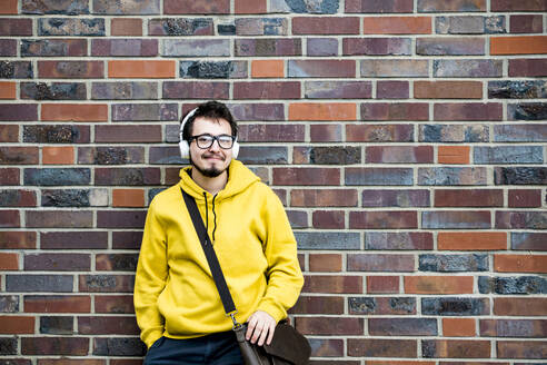 Lächelnder Student mit Tasche, der vor einer Backsteinmauer Musik hört - IHF00590