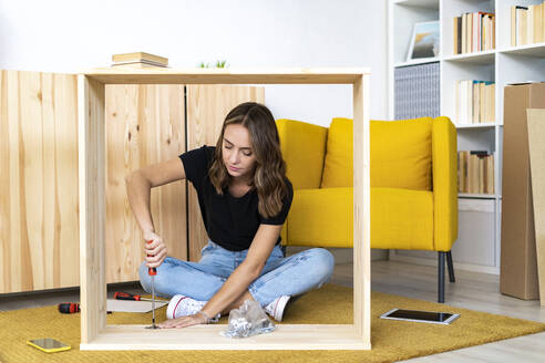 Woman assembling furniture while sitting cross-legged at home - GIOF13984