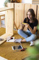 Junge Frau sitzt im Schneidersitz und benutzt ein Smartphone im Wohnzimmer - GIOF13994