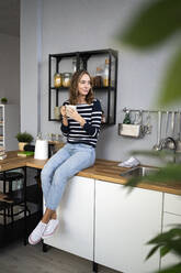 Smiling woman with mug sitting on kitchen counter - GIOF14011