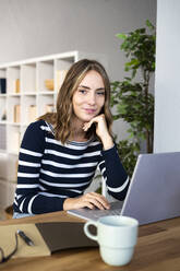 Smiling young woman with hand on chin in front of laptop at kitchen counter - GIOF14019